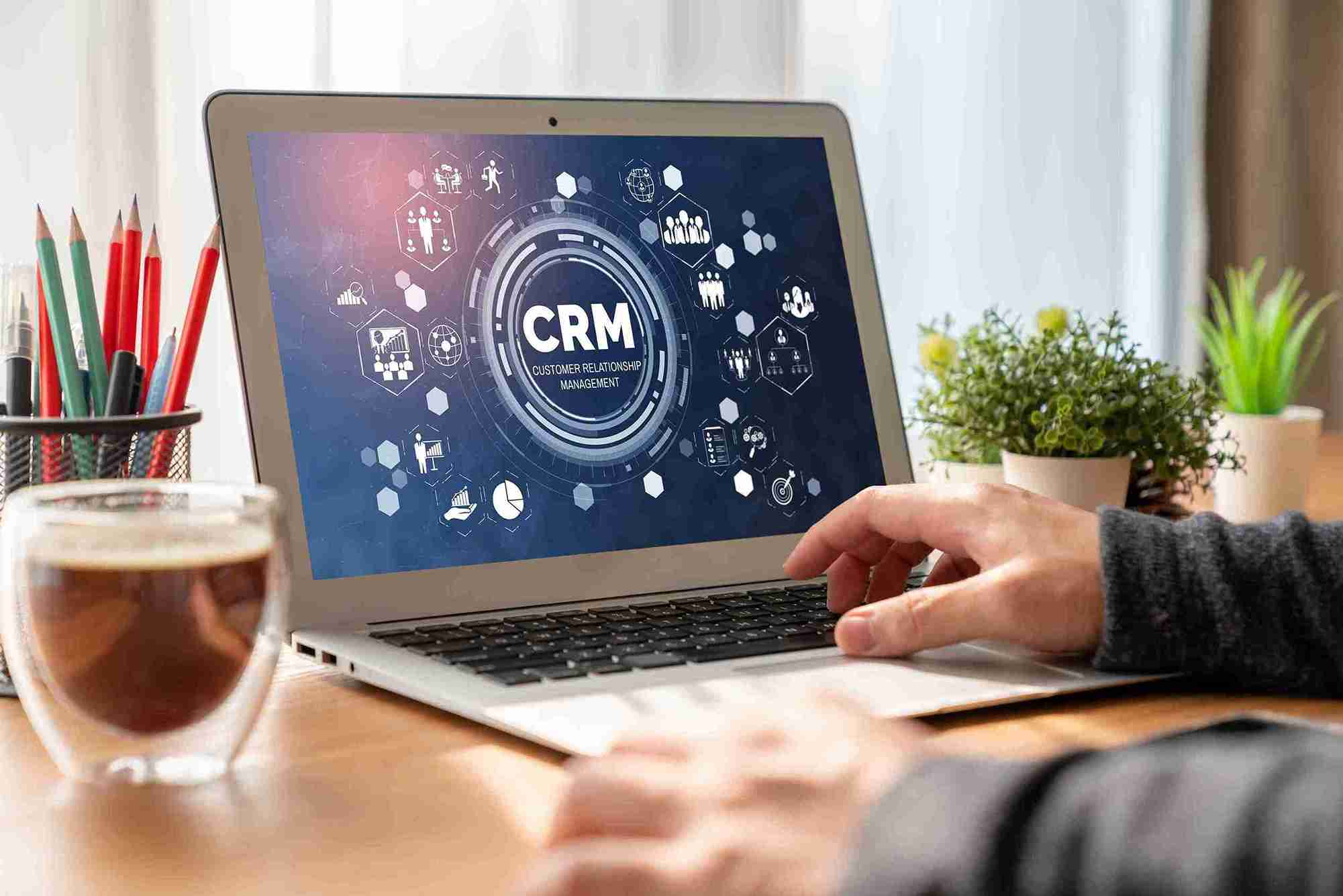 Hands typing on a laptop displaying a CRM dashboard with icons. A cup, pencils, and a plant on the wooden desk suggest a work setting.