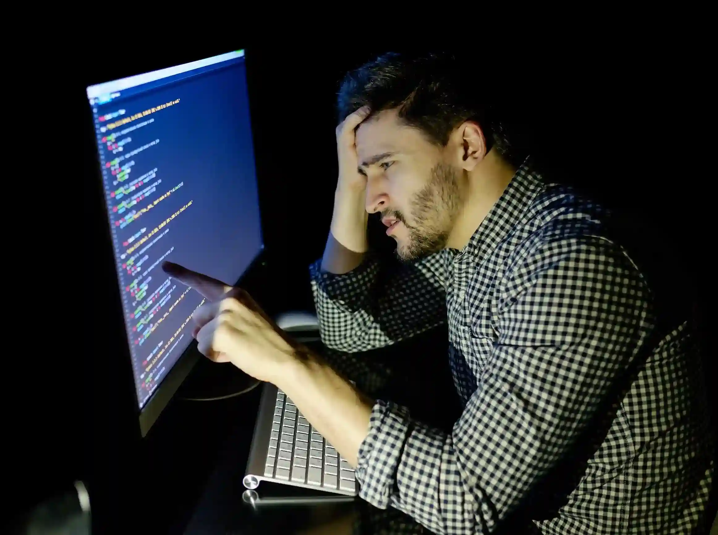 Man in plaid shirt looks frustrated, pointing at computer screen with code in dark room. His expression shows concentration and concern.