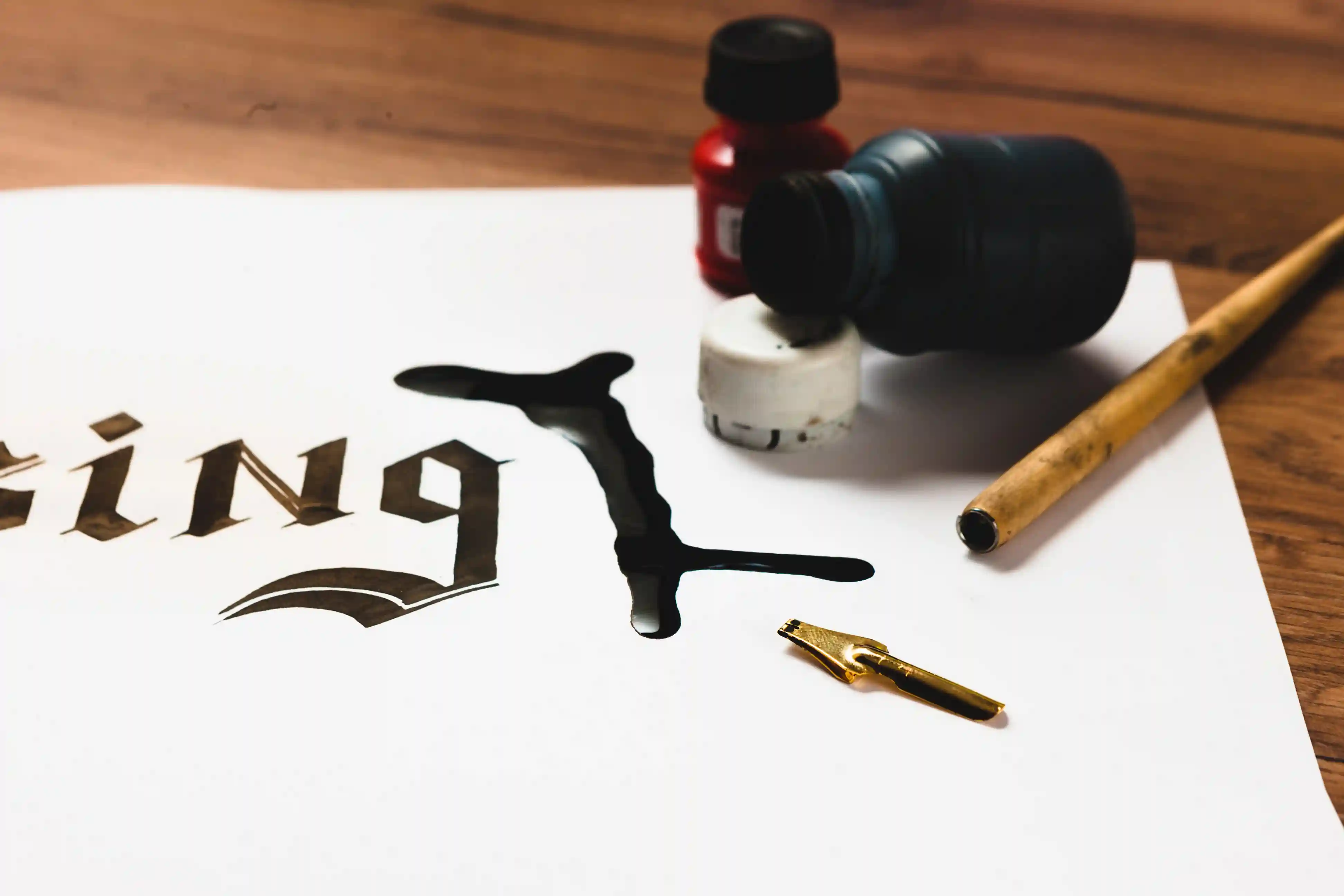 Ink spill on paper with calligraphy, red and blue ink bottles, and a gold nib pen on a wooden surface.