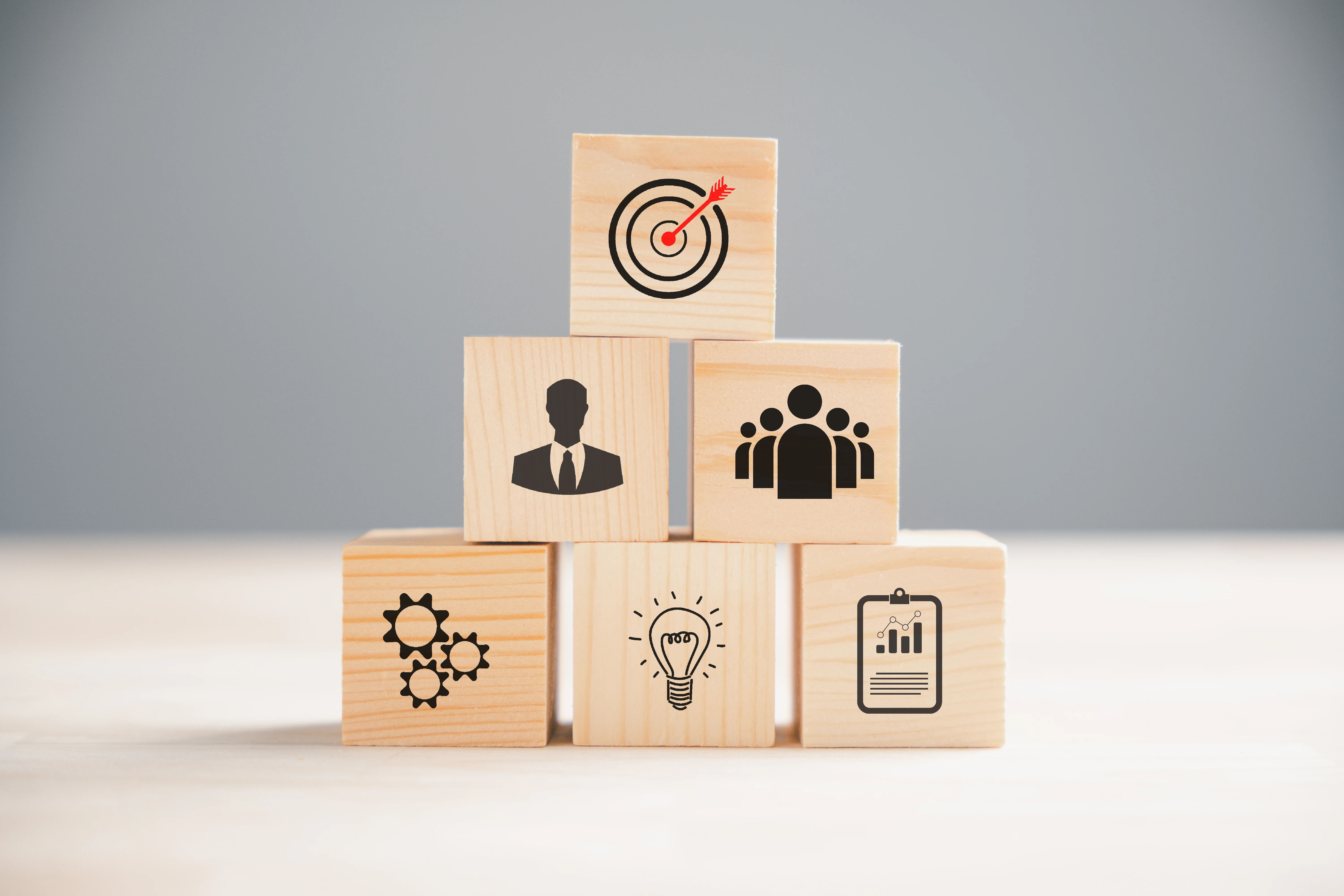 Wooden blocks with icons: target, person, group, gears, lightbulb, clipboard. Gray background, representing business concepts.