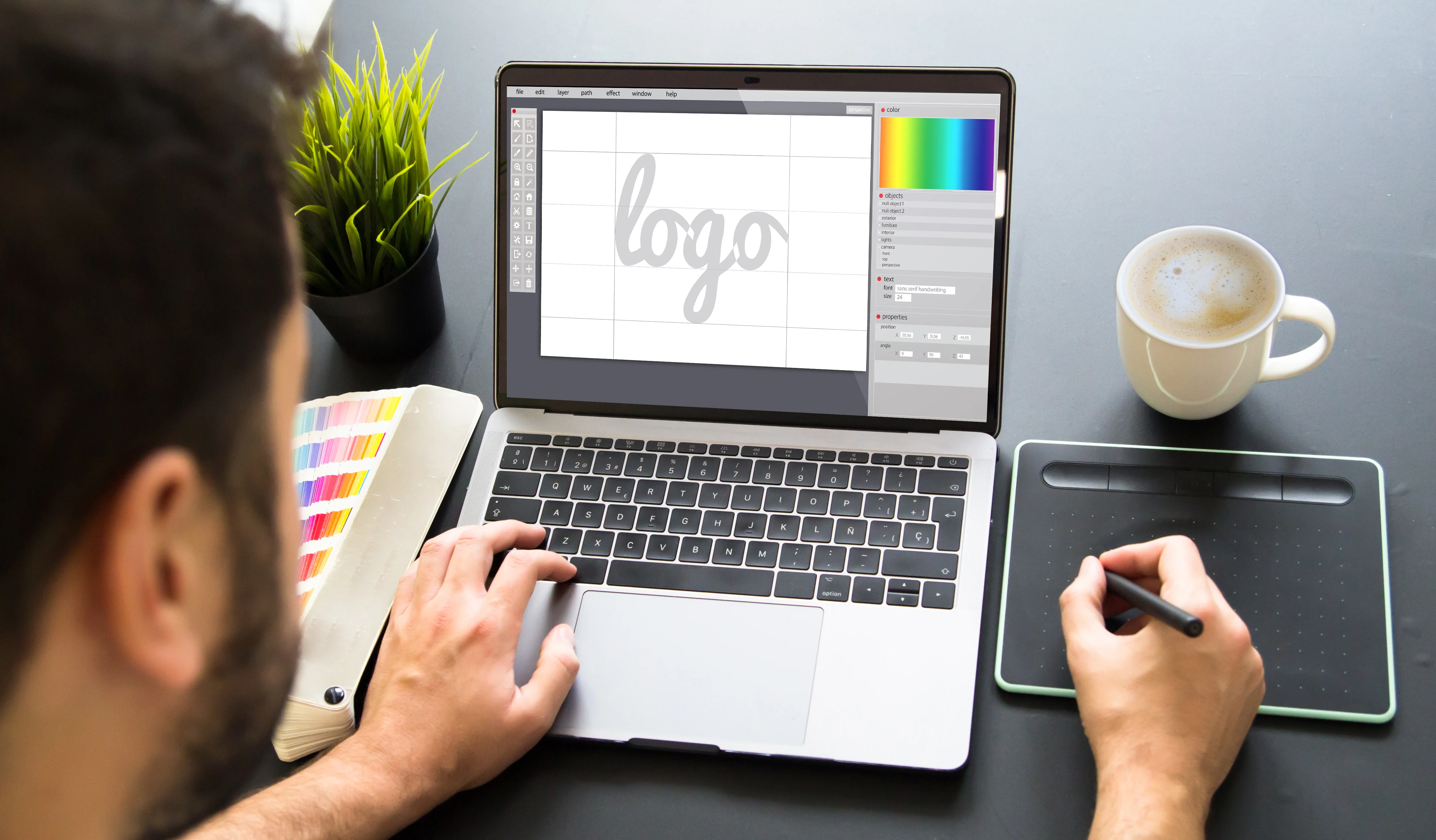 Person designs a logo on a laptop using a graphics tablet. Desk has a color palette, green plant, and coffee cup. Creative workspace vibe.
