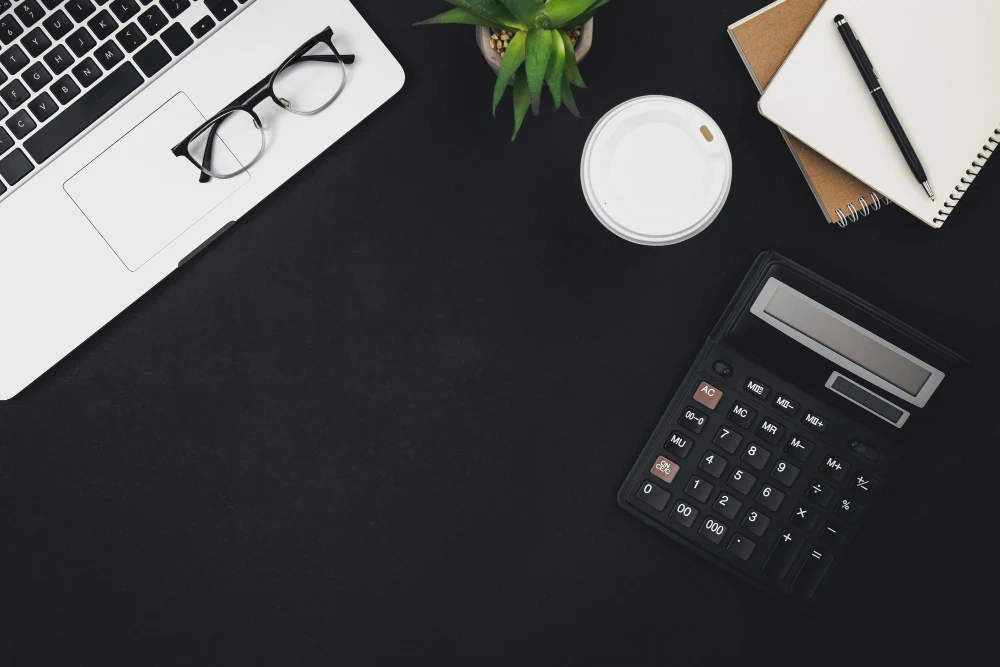 Laptop, glasses, coffee, calculator, and notebooks with pen on a black desk. Green plant adds a touch of nature. Calm, organized vibe.