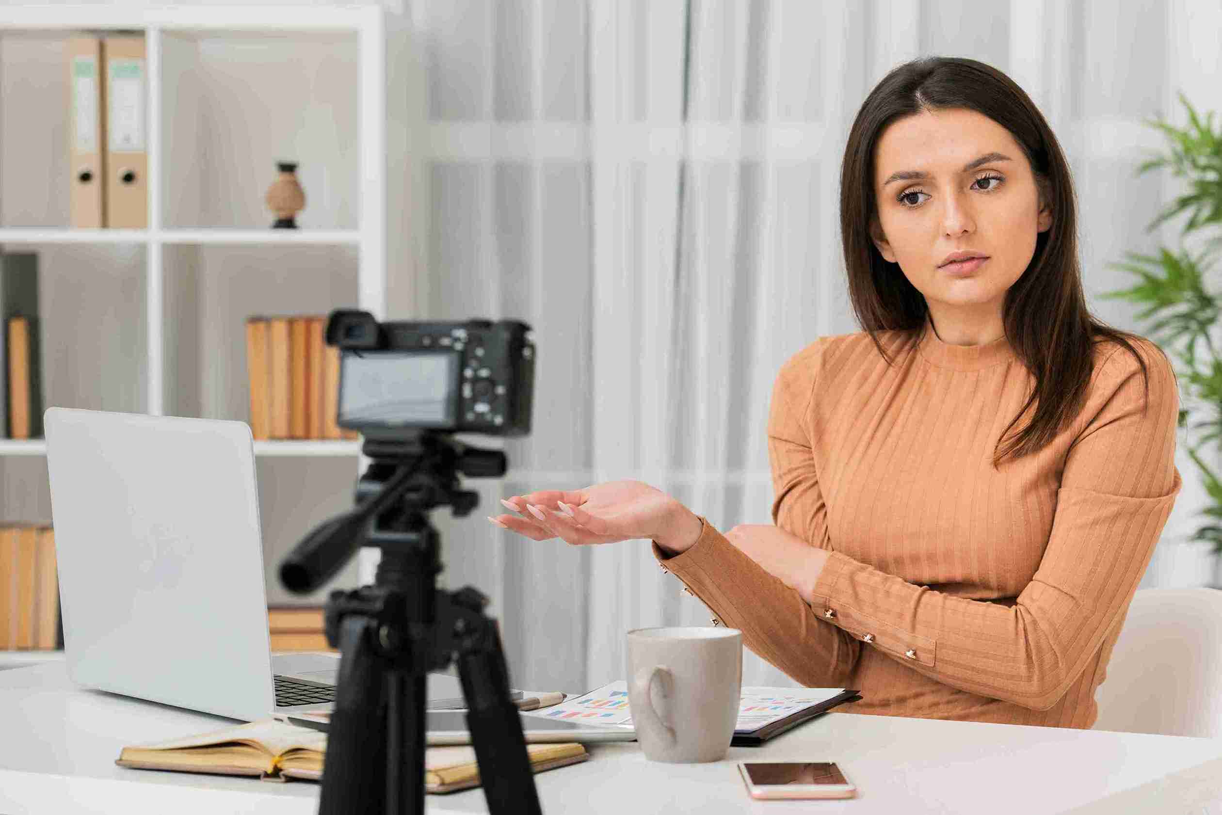 Woman in a peach top sits at a table with a laptop and open book, gesturing at a camera on a tripod. Bookshelves and plant in background.