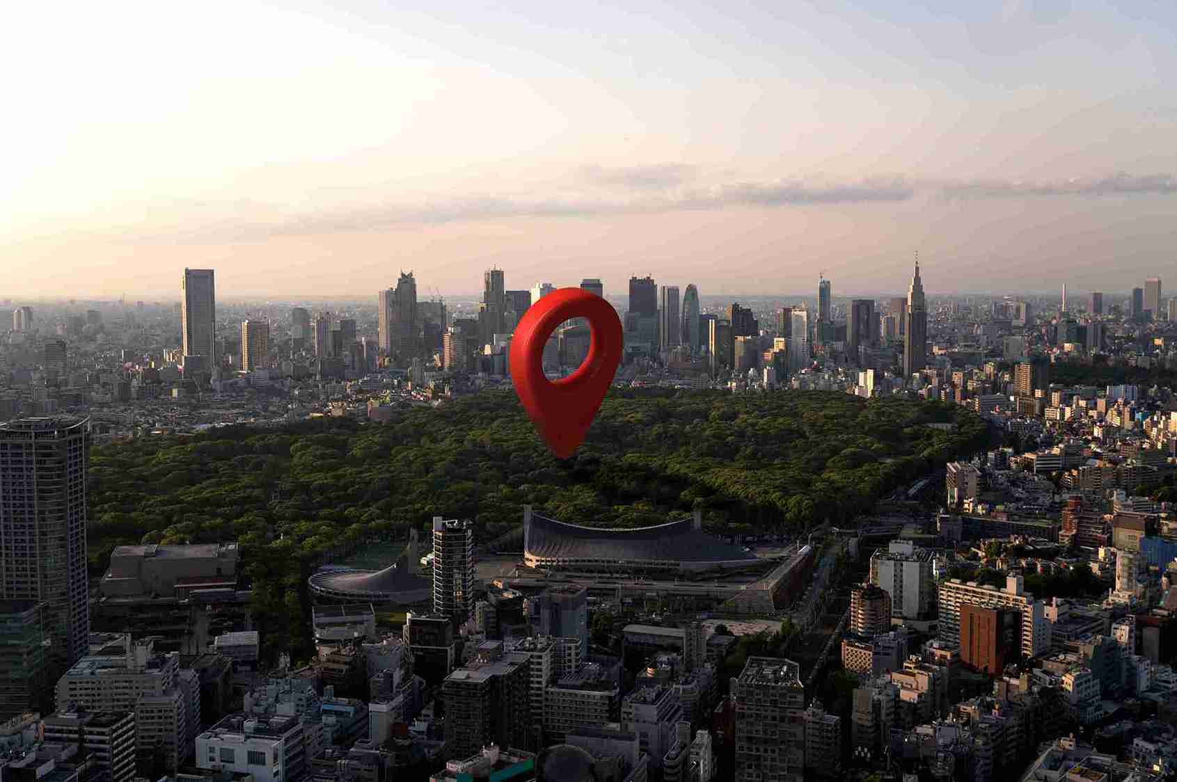 A large red location pin hovers over a cityscape with skyscrapers, green park, and stadium at sunset. Urban setting, peaceful mood.